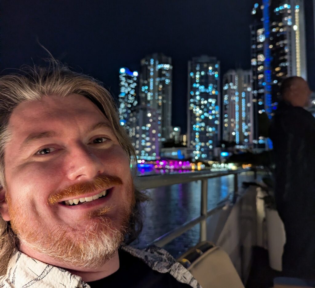 Zigarot on a boat, leaning back against a railing, smiling at the camera, with a night cityscape in the background